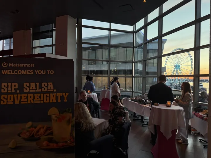 A Mattermost sign reading Sip, Salsa, Sovereignty at an evening reception with a Ferris wheel outside the windows