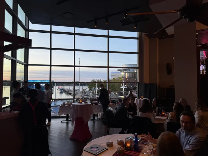 Guests at tables in front of tall windows looking out on the water at sunset
