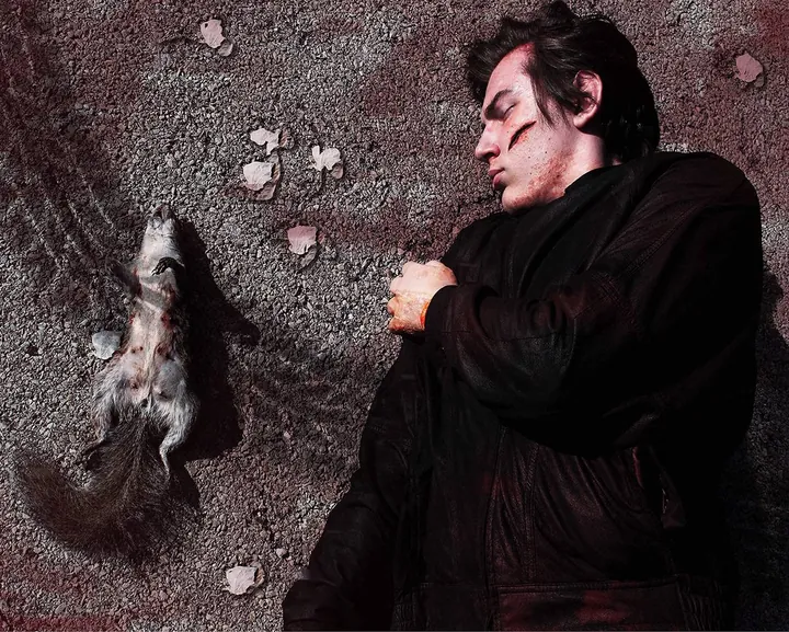 Greg lying on gravel beside a dead squirrel, eyes closed