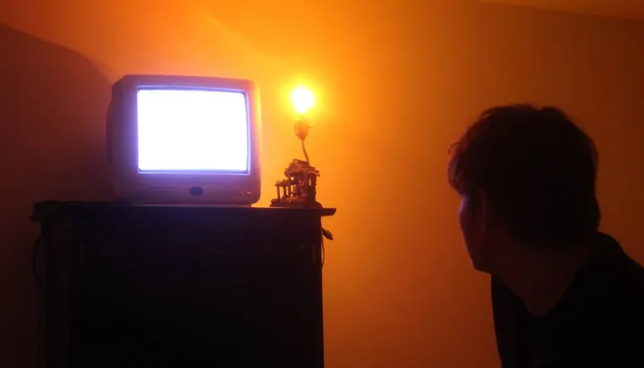 A figure in silhouette beside a glowing television in an orange room